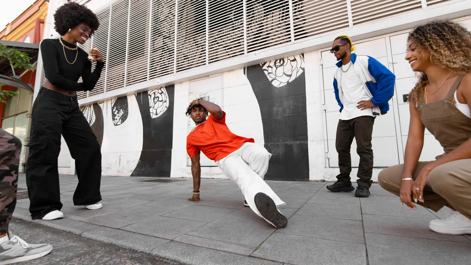 A breakdancer in an orange shirt freezes on one hand while friends cheer beside a street mural in a hip-hop scene