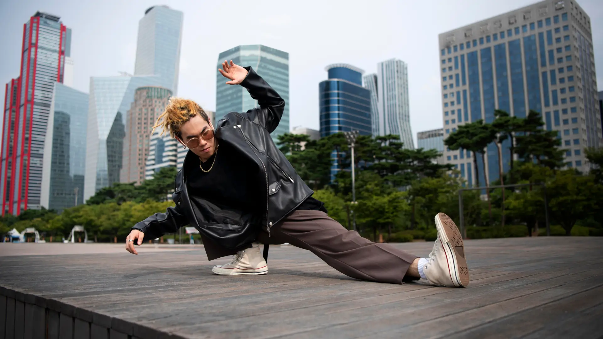 Urban hip-hop dancer in sunglasses and leather jacket striking a dynamic freeze on a wooden plaza with skyscrapers behind him