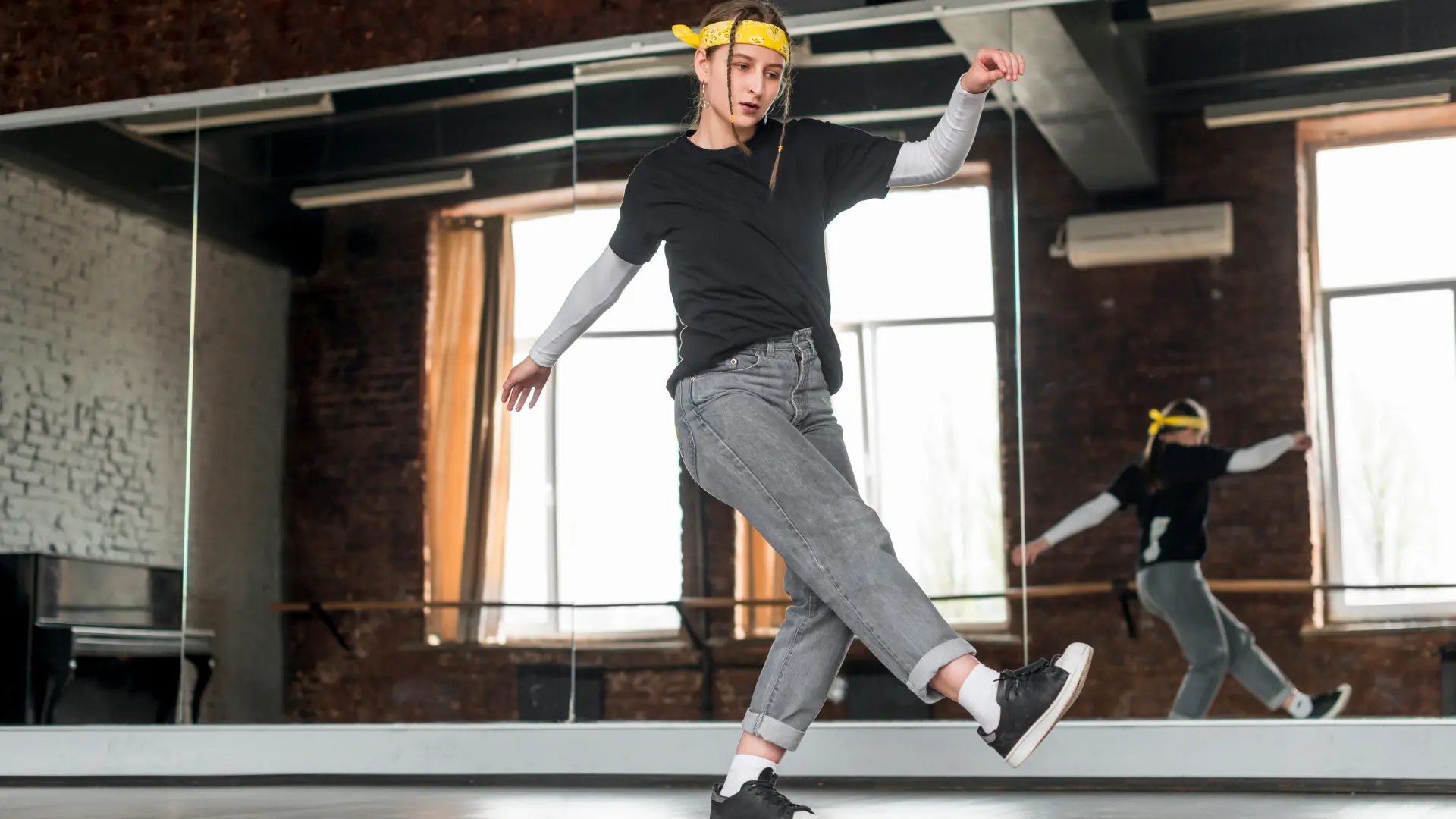 Hip-hop dancer wearing a yellow bandana, black tee and rolled grey jeans practicing choreography in a mirrored studio