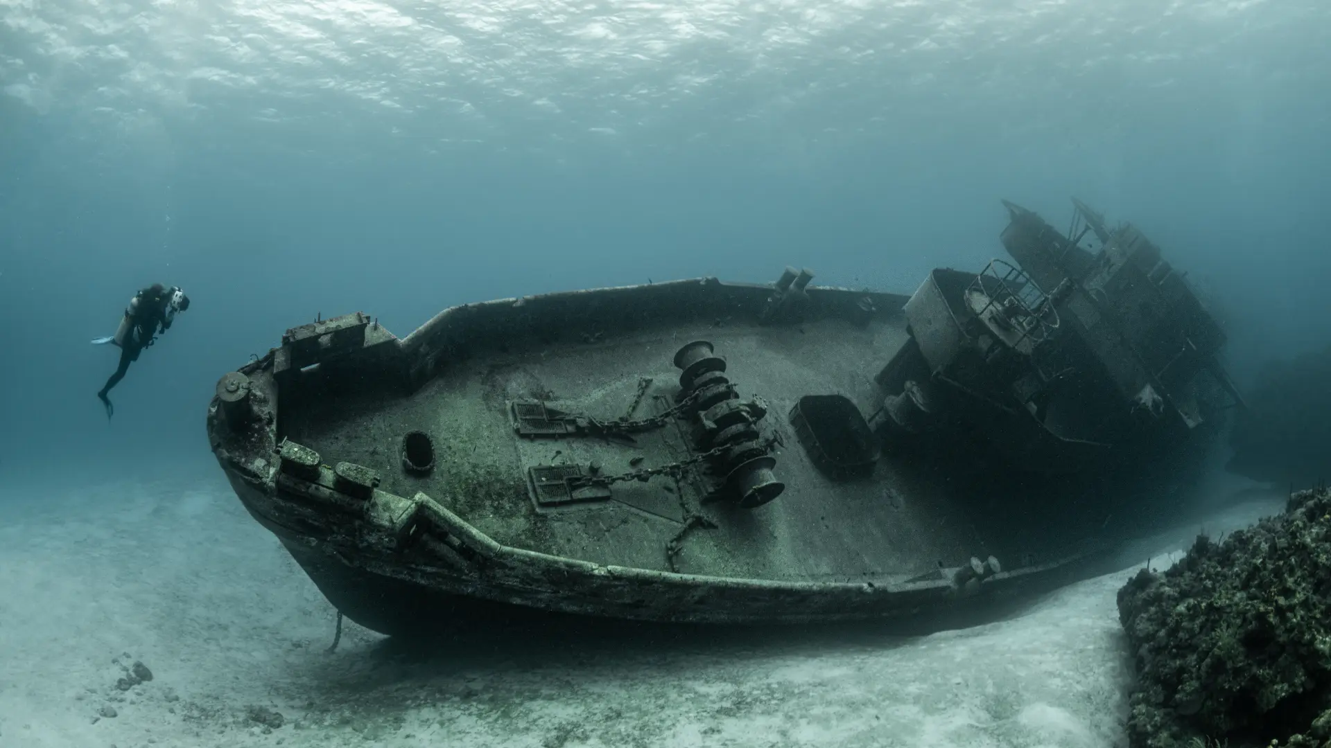 Diver explores rusted shipwreck on sandy seabed, broken deck and hull covered in marine growth under deep blue water