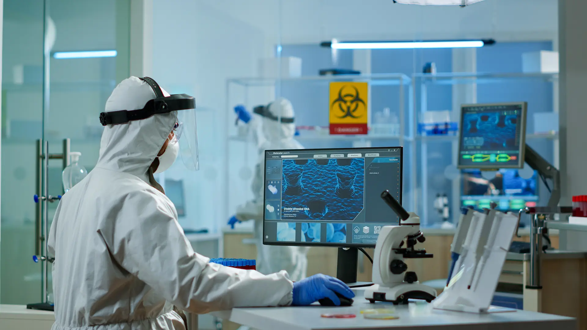 Lab technician in full PPE studies DNA imaging on a computer in a biohazard lab, showing radiation applications in healthcare