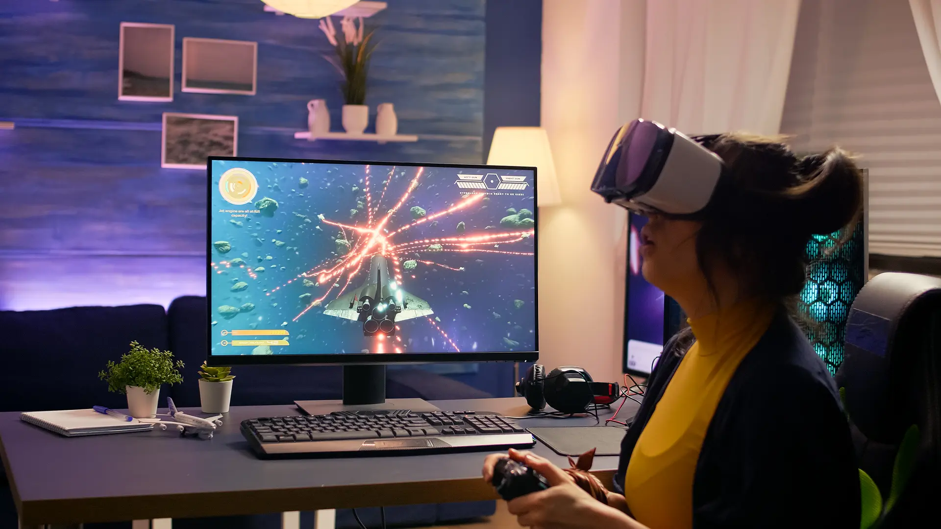 Woman in VR headset plays a polygonal space shooter on a PC monitor with keyboard, controller and blue ambient room lighting